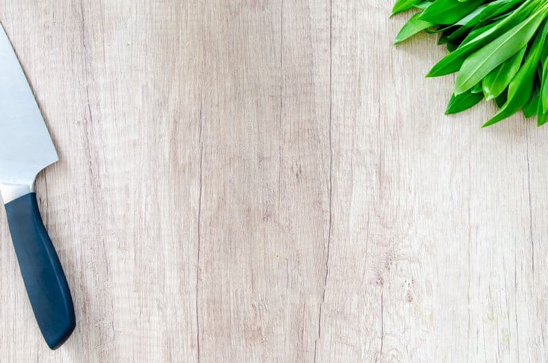 A kitchen knife resting on a wooden cutting board next to fresh greens