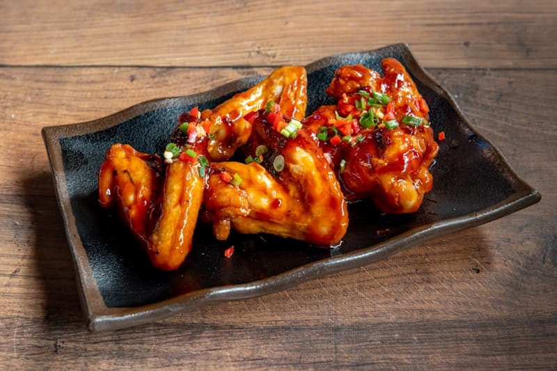 Glossy red spicy grilled chicken wings on a black platter, garnished with sesame seeds and scallion