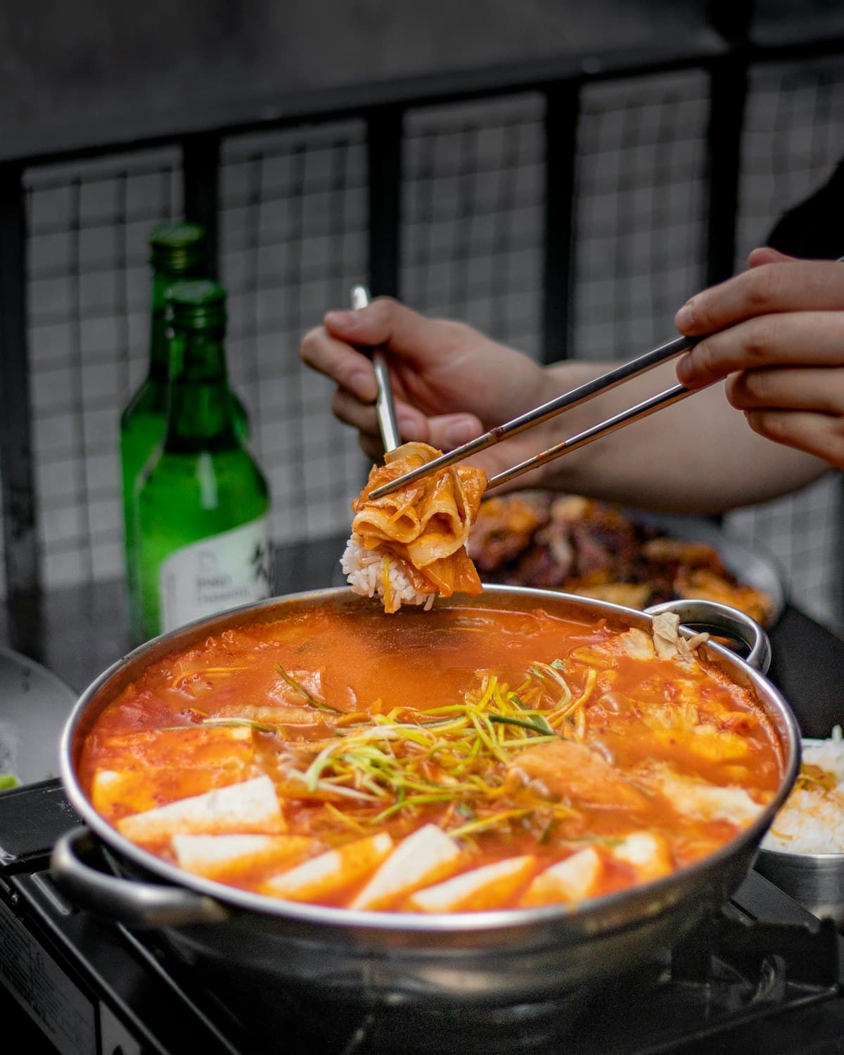 Kimchi jjigae with tofu in a bubbling metal pot, kimchi lifted with chopsticks over rice