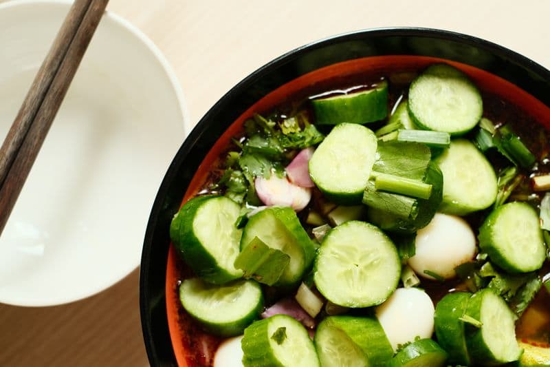 Cold cucumber soup with sliced cucumber, herbs, and quail eggs in a dark bowl