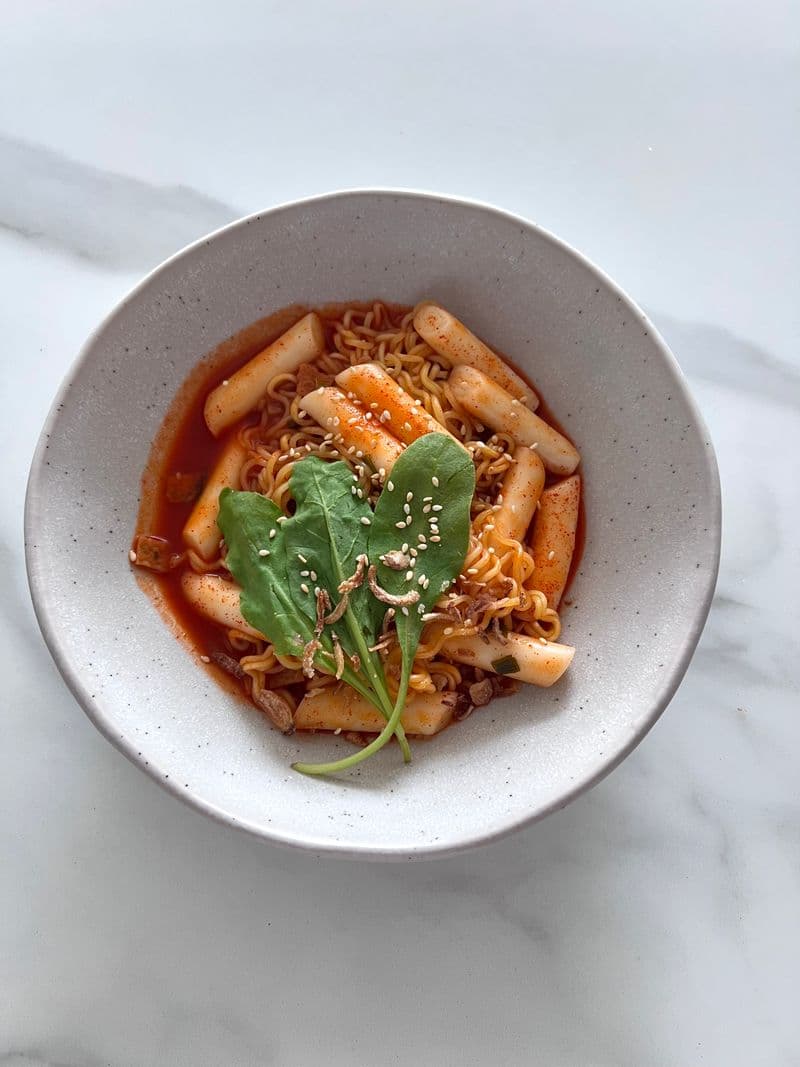 Rice cakes and ramen noodles in red sauce topped with sesame seeds in a bowl