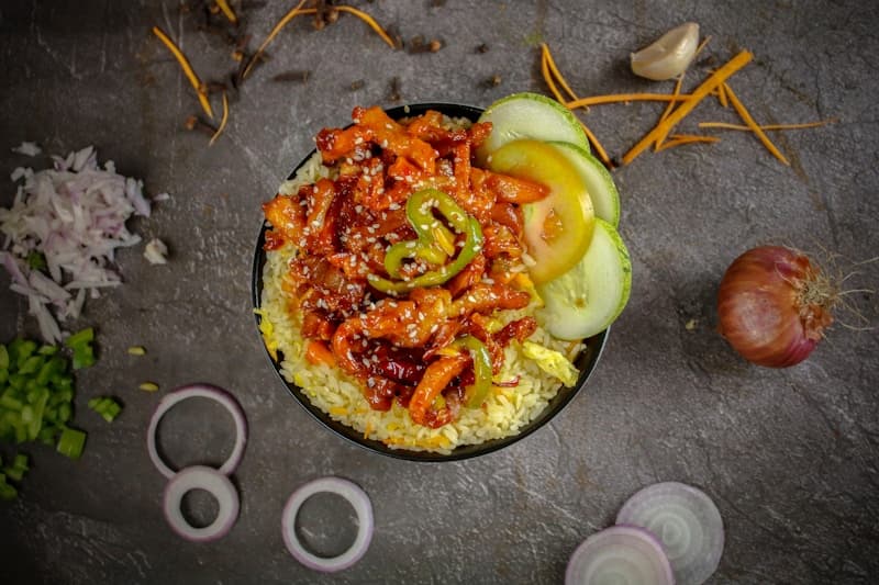Spicy stir-fried pork over rice with cucumber slices in a black bowl