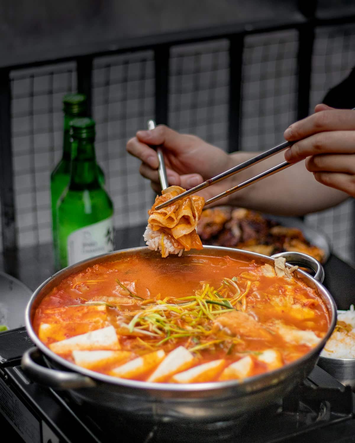 Kimchi jjigae with tofu in a bubbling metal pot, kimchi lifted with chopsticks over rice