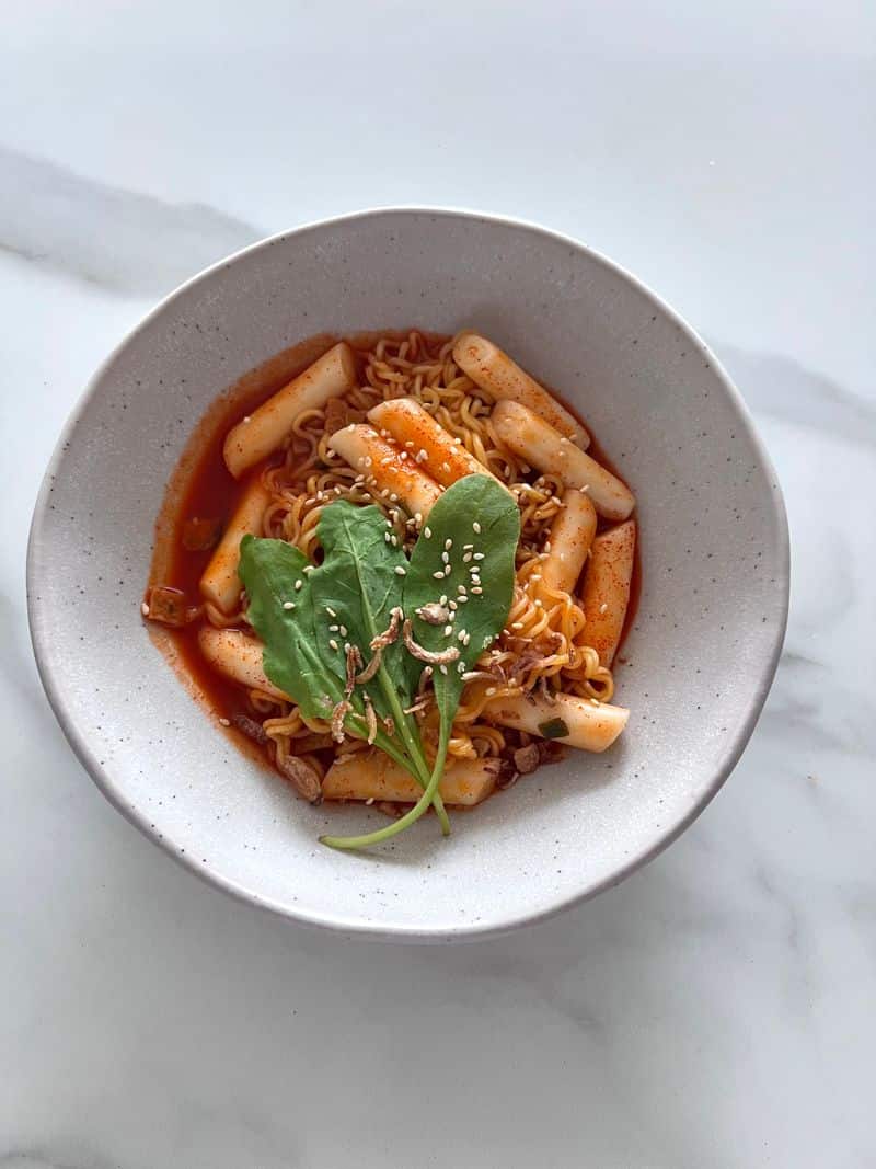 Rice cakes and ramen noodles in red sauce topped with sesame seeds in a bowl