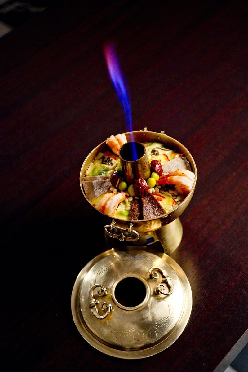 Brass hot pot with a central chimney and lit flame, filled with prawns, beef, jujube and ginkgo nuts